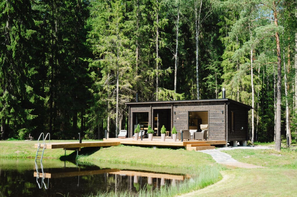 Moderni puinen mökki, jossa on terassi, sijaitsee pienen lammen rannalla, jota ympäröivät korkeat vihreät puut metsäisellä alueella.