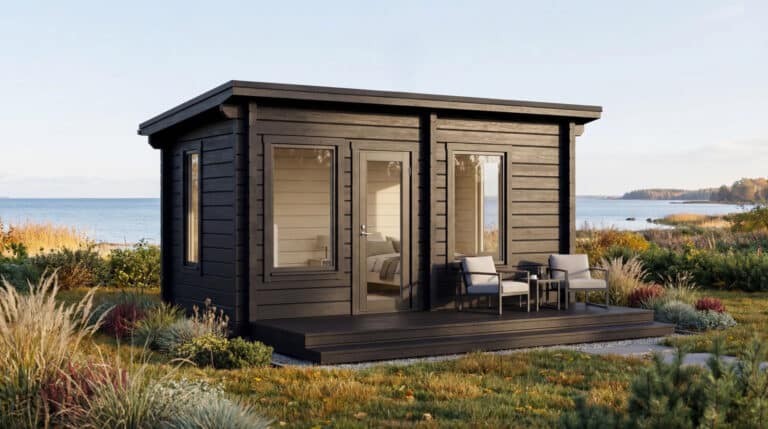 Pieni, moderni musta mökki, jossa on suuret ikkunat ja kannella, jossa on kaksi tuolia, sijaitsee nurmikolla lähellä vettä, ja taustalla on rannikkomaisema.