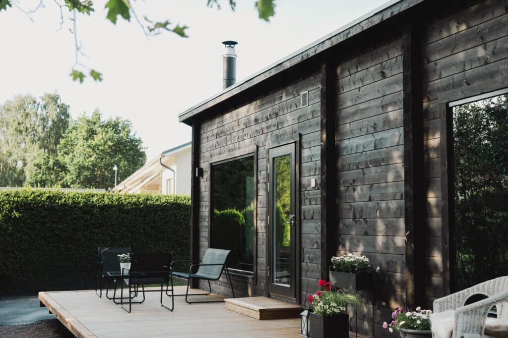 Moderni tummaa puuta oleva Saunamökki, jossa on suuret ikkunat, savupiippu ja puinen kansi, jossa on mustat ulkokalusteet ja ruukkukukkia, vehreyden ympäröimänä.