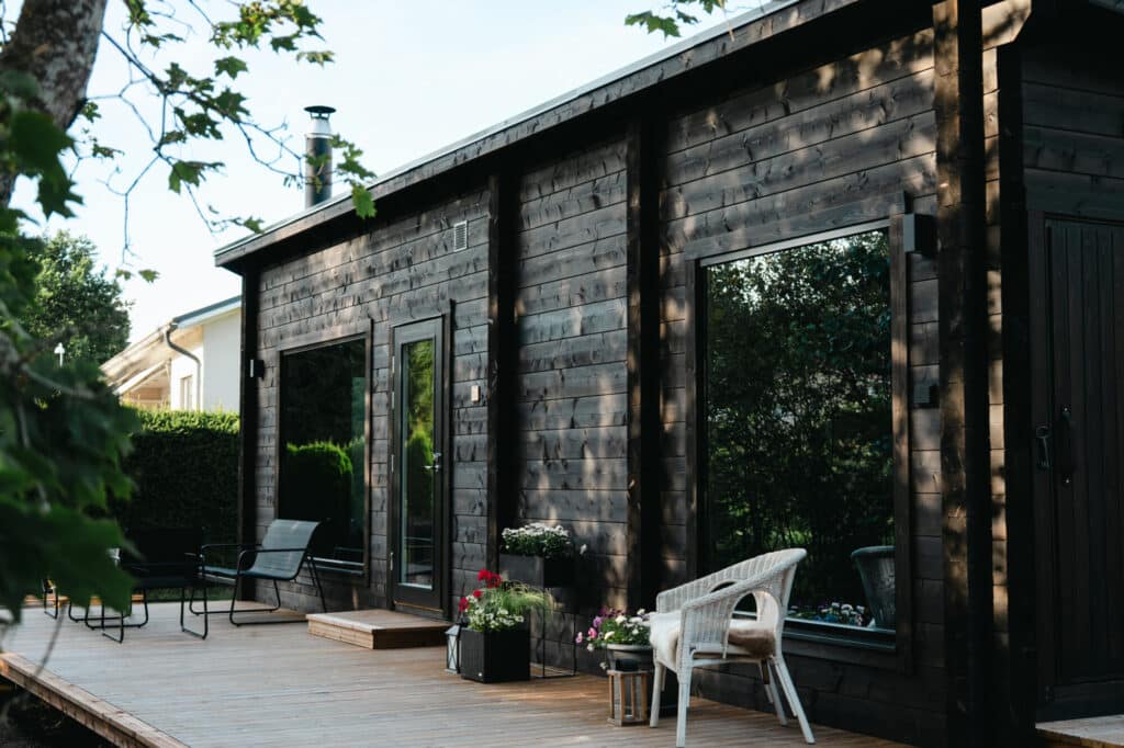 Moderni tummapuinen saunamökki, jossa on suuret ikkunat ja puinen kansi, joka on kalustettu ulkotuoleilla, pöydillä ja ruukkukasveilla.