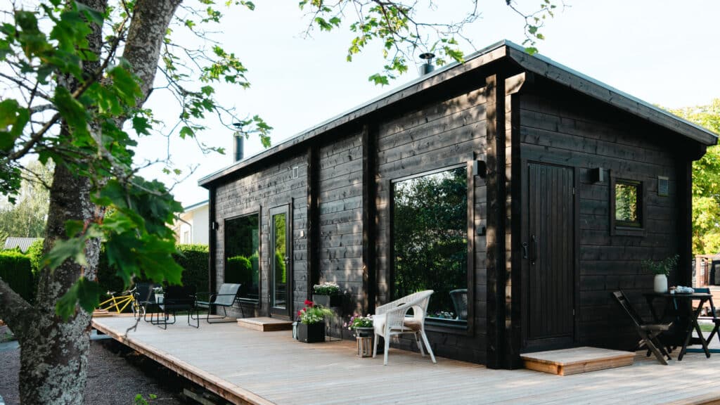 Ehtan moderni, tumma hirrestä valmistettu valmis saunamökki, jossa on maisemaikkunat ja puinen terassi. Terassilla on ulkokalusteita ja edustalla kasvaa iso vaahtera.