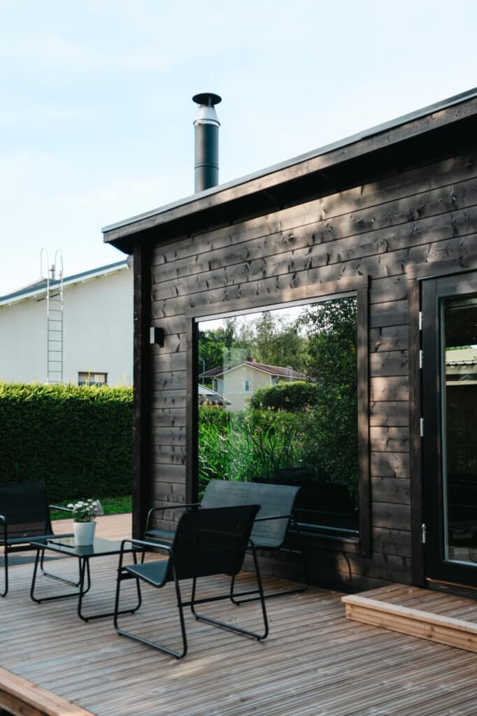 Moderni puinen kansi, jossa on mustia tuoleja ja pöytä, istuu tummanpuisen saunamökin vieressä; taustalla näkyvät tikkaat ja rehevä vehreys.