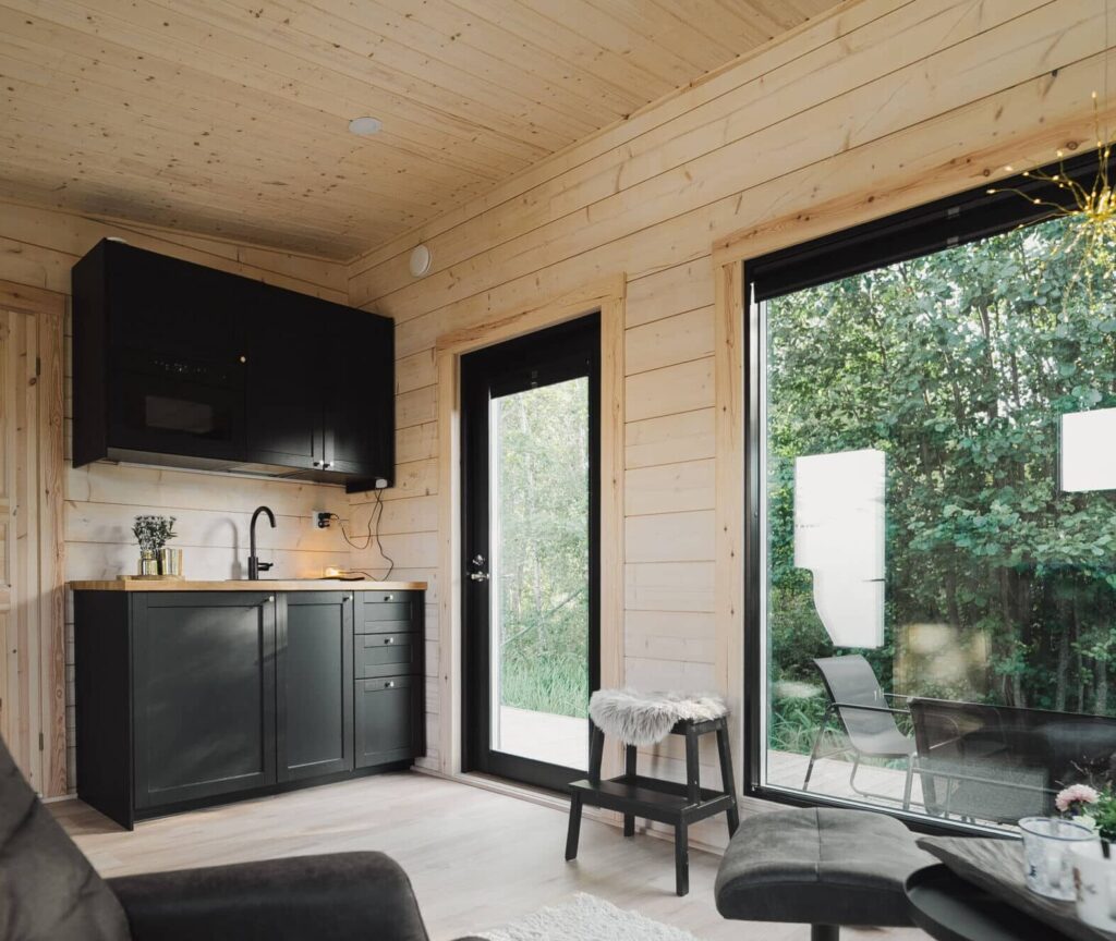 Kompakti moderni keittiö Ehta Saunamökki 30:ssä, jossa on mustat kaapit, vaaleat hirsiseinät ja katto, lasiovet ja maisemaikkuna,