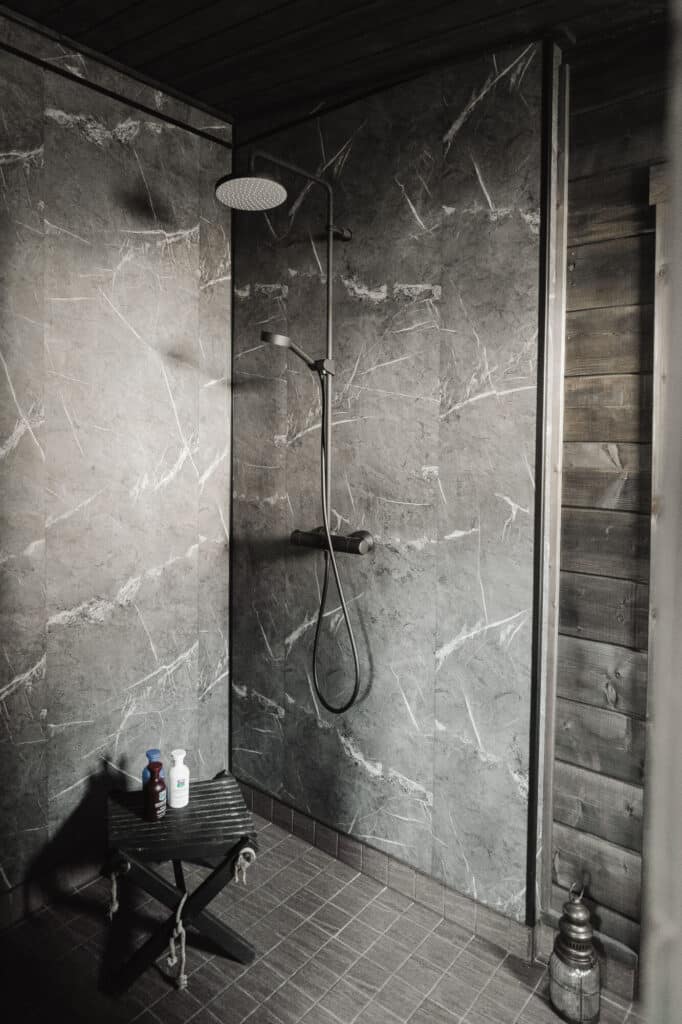 Moderni suihku, jonka seinät ovat harmaata marmoria, sadesuihkupää ja puinen jakkara, jossa on pulloja, seisoo puupaneloitujen seinien vieressä luoden rentouttavan Saunamökki 30 -inspiroituneen retriitin.