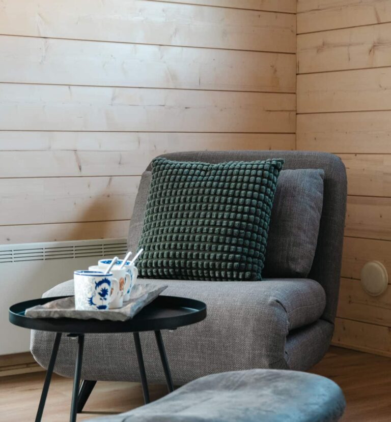 Kotoisa harmaa nojatuoli, jossa on kuvioitu vihreä tyyny, istuu pienen pyöreän mustan pöydän vieressä, jossa on kaksi mukia vaaleassa hirrestä valmistetussa saunamökissä.