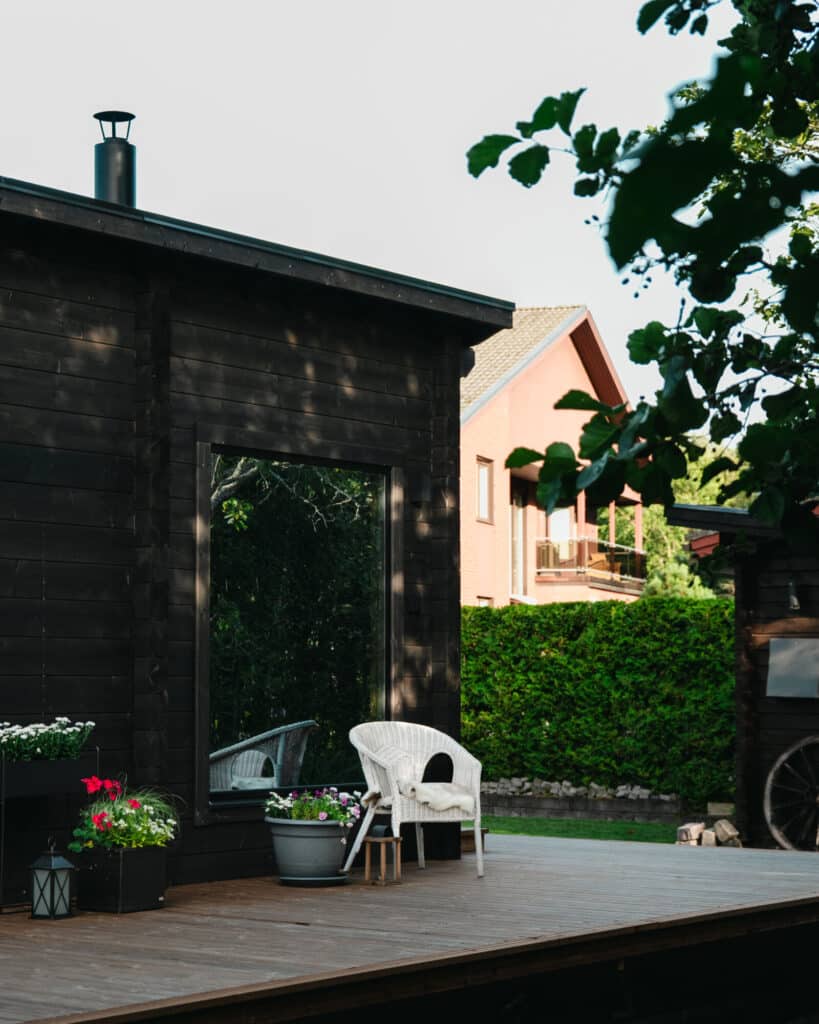 Valkoinen tuoli ja ruukkukukkia istuu tumman Saunamökin puisella kannella, taustalla näkyy vehreyttä ja toinen talo.