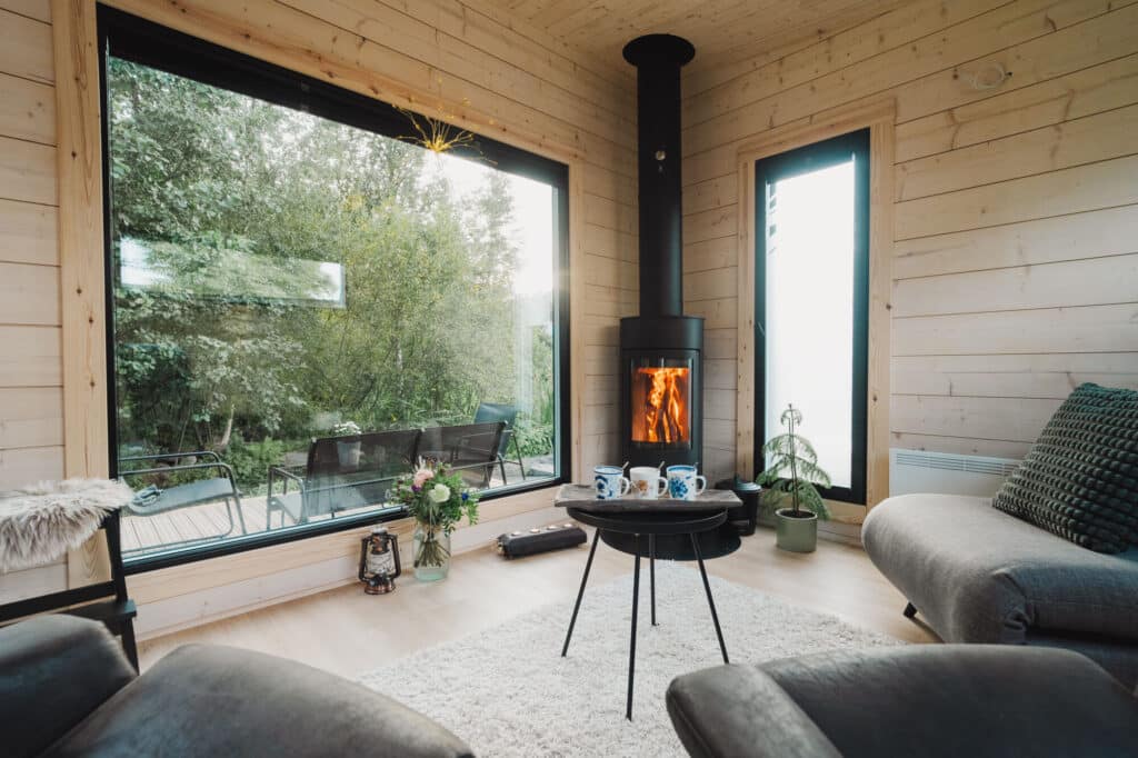 Moderni Saunamökin olohuone, jossa on vaaleat puuseinät, suuret ikkunat, musta puuhella, jossa on tuli, pyöreä sohvapöytä, jossa on mukeja ja kasveja viihtyisän sohvan vieressä.