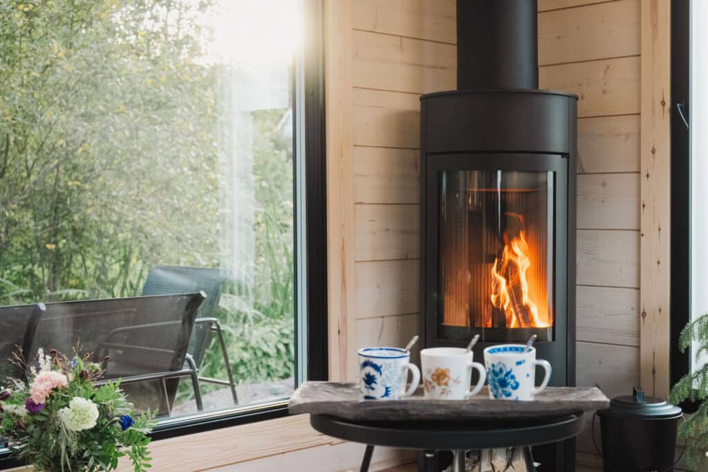 Moderni puuhella, jossa on tuli sisällä, ikkunan vieressä, josta on näkymät terassille; pöydän edessä on kukkamukeja, teekannu ja kukka-asetelma - täydellinen kodikkaisiin hetkiin Saunamökki 30:ssä.