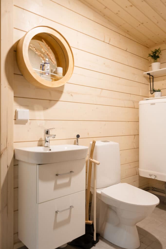 Pieni valmis kylpyhuone, jossa on vaaleat hirsiseinät, valkoinen pesuallas alalaatikoilla, wc-istuin sekä pyöreä Tapio Anttilan puukehikolla kehystetty peilihylly.