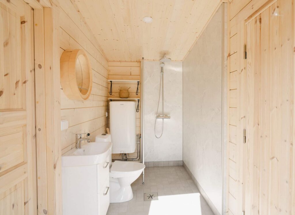 Ehta valmis kylpyhuone, jossa on vaaleat hirsiseinät, valkoinen pesuallas, wc, moderni suihkutila sadesuihkulla ja lasiseinällä sekä pyöreä design-peilihylly pesualtaan yläpuolella.