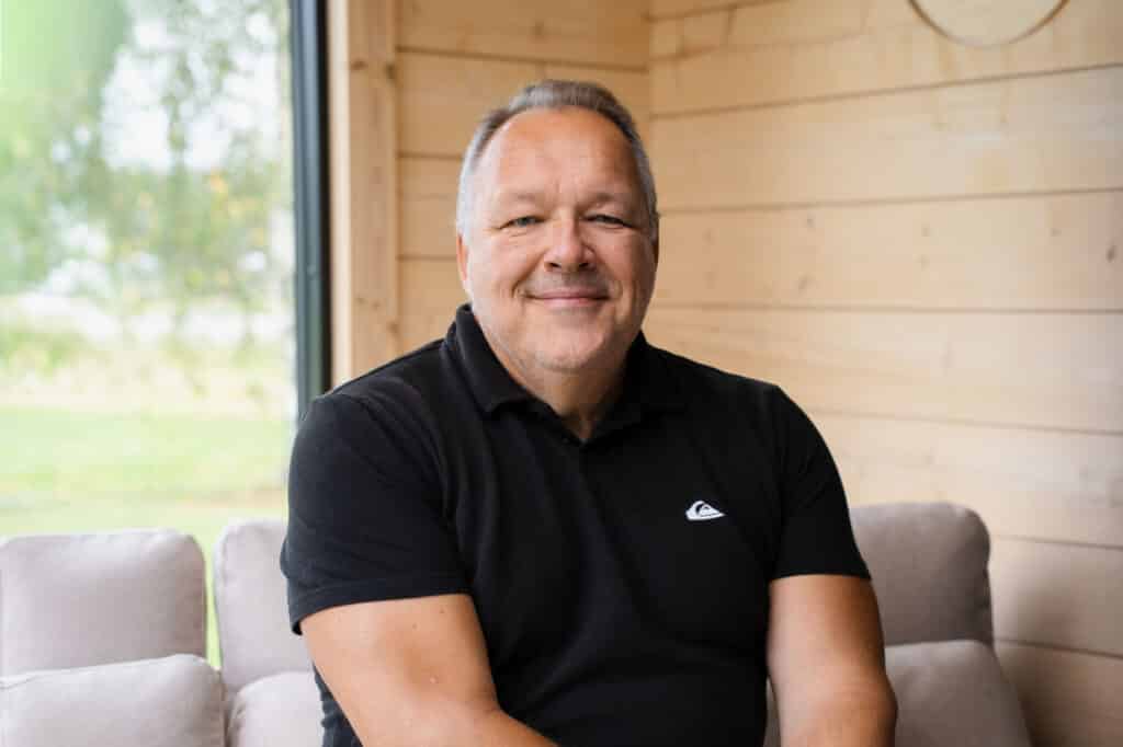 Mies, jolla on yllään musta poolopaita, istuu vaalealla sohvalla puuhuoneessa, jonka ikkunasta avautuu näkymä ulkona olevaan vehreään maisemaan.