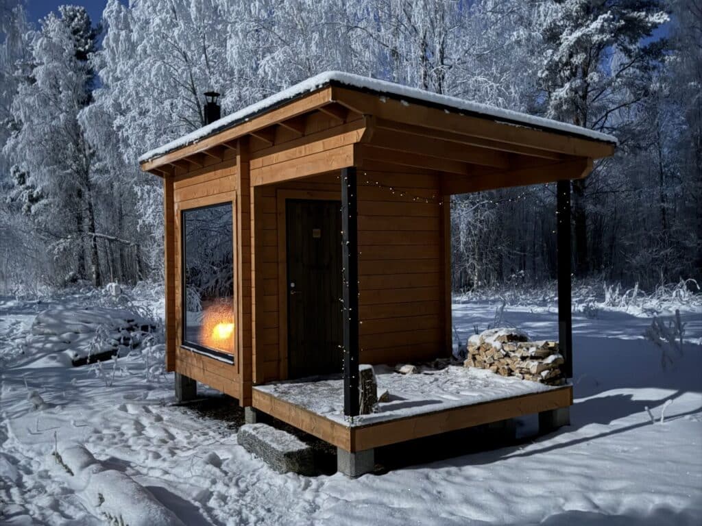 Pieni puinen saunarakennus, jossa on suuri ikkuna ja kuisti, ja jota ympäröivät lumipeitteiset puut.
