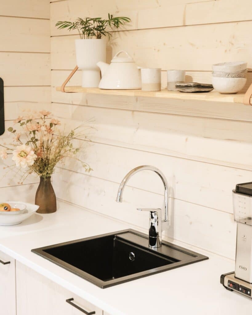 Moderni hirsimökin keittiö, jossa on musta pesuallas, kromihana, valkoinen työtaso, puinen astiahylly ja kukkamaljakko vaalealla puupaneeliseinällä.