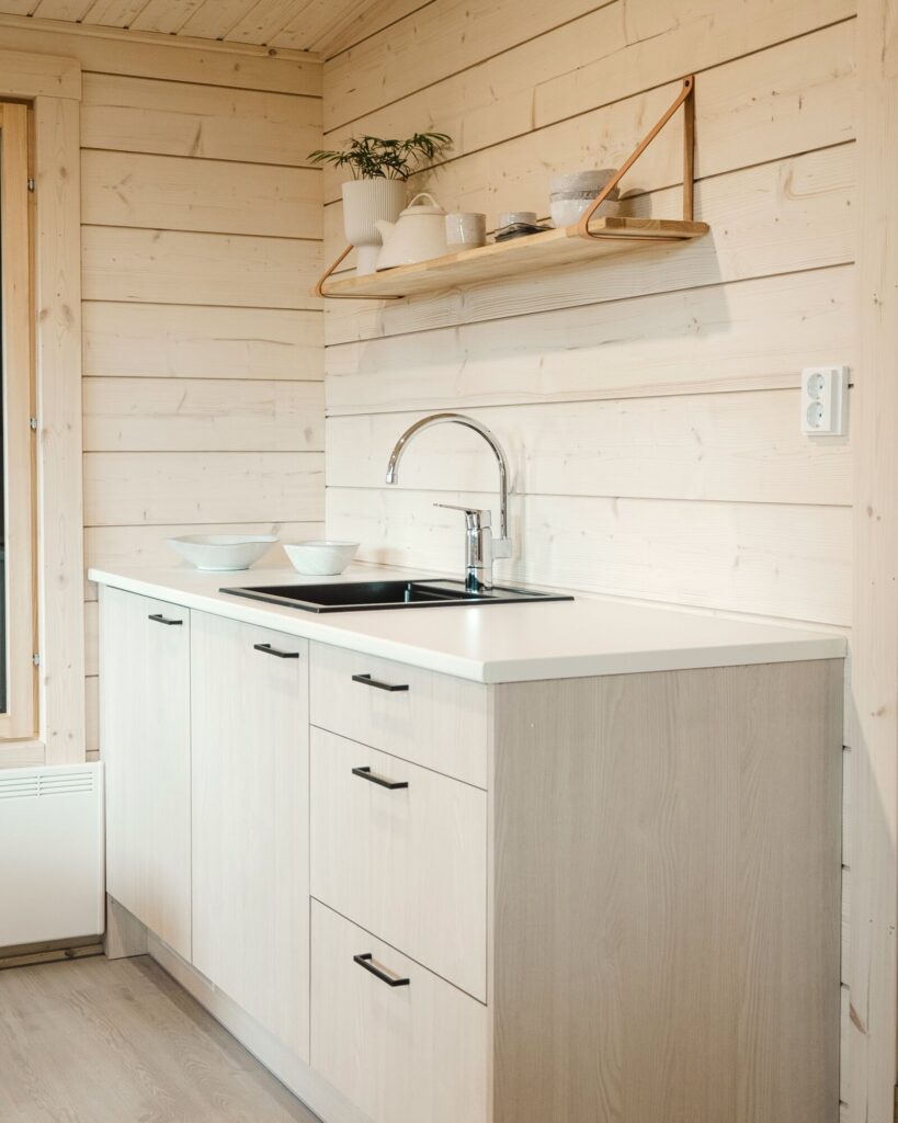 Kompakti hirsimökin keittiö, jossa on vaaleat puukaapit, musta pesuallas, kromihana, seinähylly, jossa on kasveja ja astioita, ja vaaleat puupaneeloidut seinät.