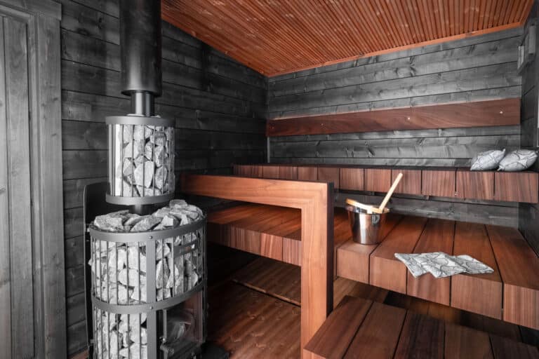 Moderni 30 neliön saunamökki, jossa on puinen saunan sisustus, porrastetut lauteet, kivillä täytetty metallinen kiuas, kauha kauhalla, Unelma-varustelu ja kuviolliset tyynyt lauteilla.