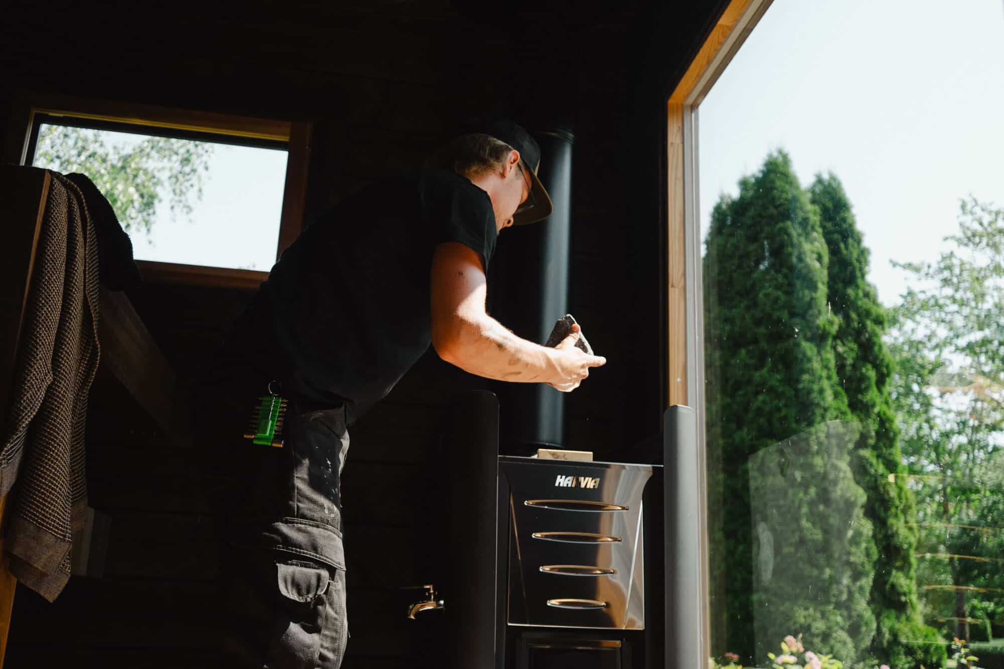 Ehta huoltomies vaihtaa kiukaaseen kiviä pihasaunassa, jonka suurista ikkunoista näkyy puita ulkona.
