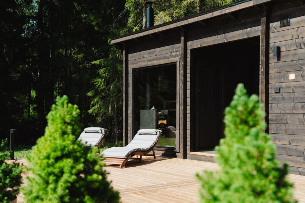 Kaksi pehmustettua lepotuolia istuu puisella kannella modernin tummasta puusta valmistetun mökin ulkopuolella, ja taustalla on vihreitä puita.
