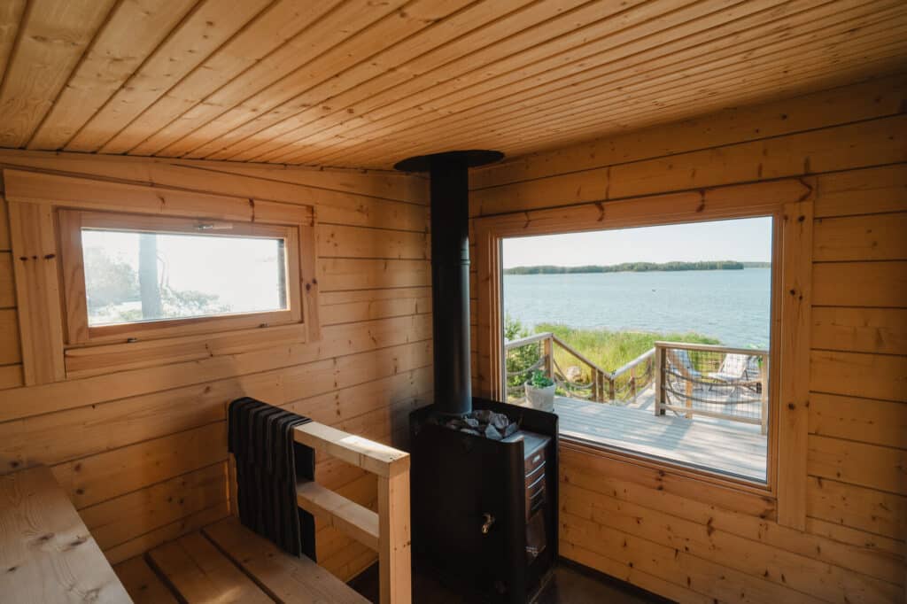 Puisen saunan sisätilat, jossa on musta kiuas, kaksi ikkunaa, joista avautuu näkymä järvelle, terassi ja viheriö ulkona.