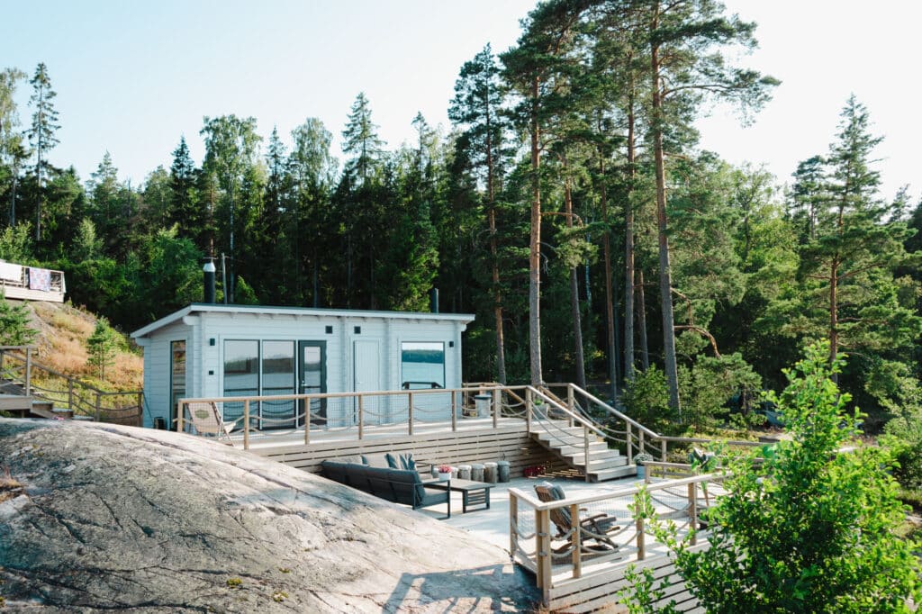 Pieni moderni mökki, jossa on suuret ikkunat, sijaitsee puisella kannella mäntyjen ja kallioisen maaston ympäröimänä metsäisellä alueella.