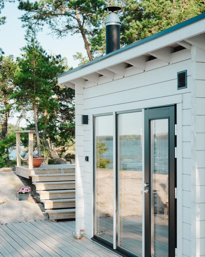 Valkoinen puinen mökki, jossa on lasiovet ja terassi, puiden ympäröimä ja josta on näkymät vesistölle aurinkoisena päivänä.