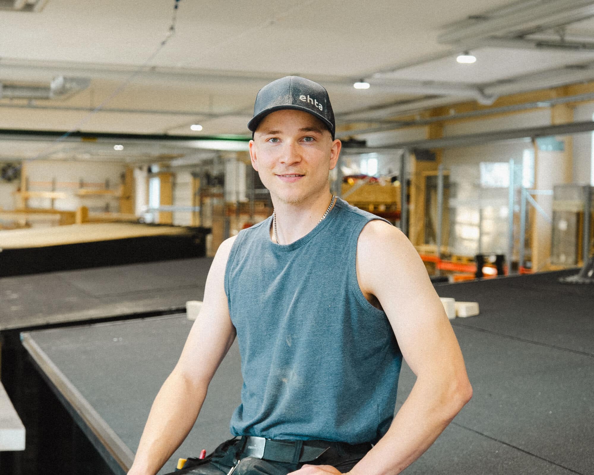 Tumma lippalakki ja hihaton paita päällään oleva saunanrakentaja seisoo rakennustyömaalla sisätiloissa, työkalut vyöllään ja rakennusmateriaalit taustalla.