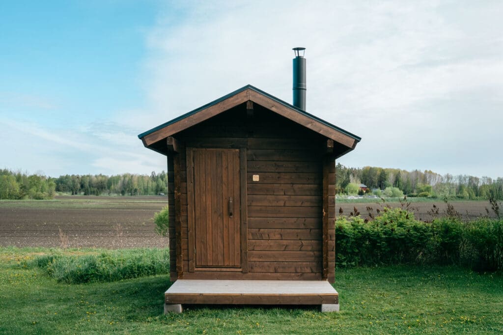 Pieni puinen pihasaunamökki, jossa on metallinen savupiippu, istuu nurmikolla, taustalla peltoja ja puita osittain pilvisen taivaan alla lähellä Marketanpuistoa.