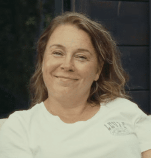 Kameraan hymyilee nainen, jolla on aaltoilevat ruskeat hiukset, valkoinen Levis-t-paita yllään ja joka istuu sisätiloissa tummalla taustalla valmiina Ehtan talvikampanjaan 2025.