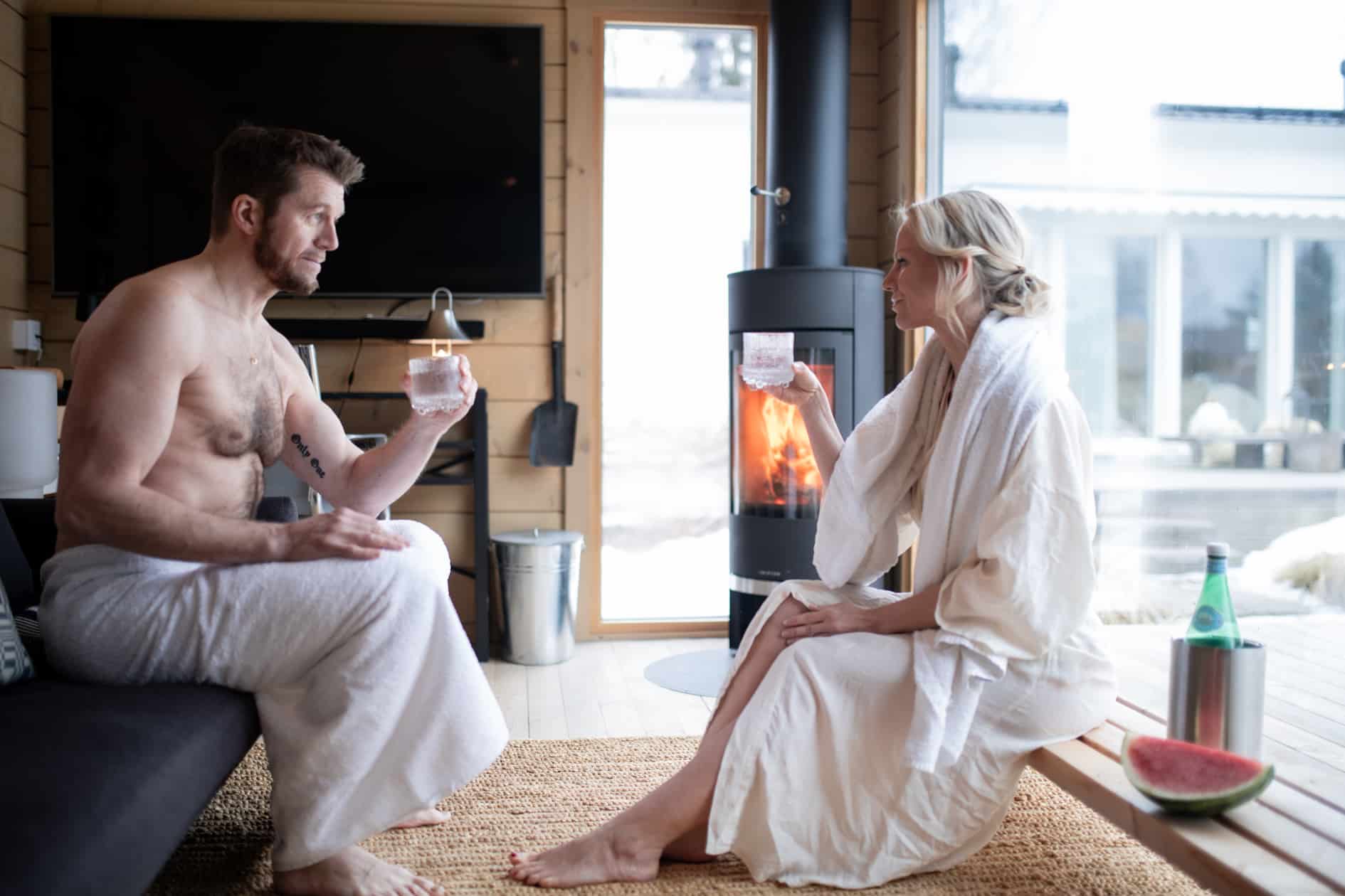 Pyyhkeisiin pukeutuneet mies ja nainen istuvat Ehta saunamökissä takan äärellä nauttimassa juomia. Pöydän vieressä on vesipullo ja vesimeloniviipaleita. Suurista ikkunoista näkyy lunta - täydellinen paikka rentoutumiseen.