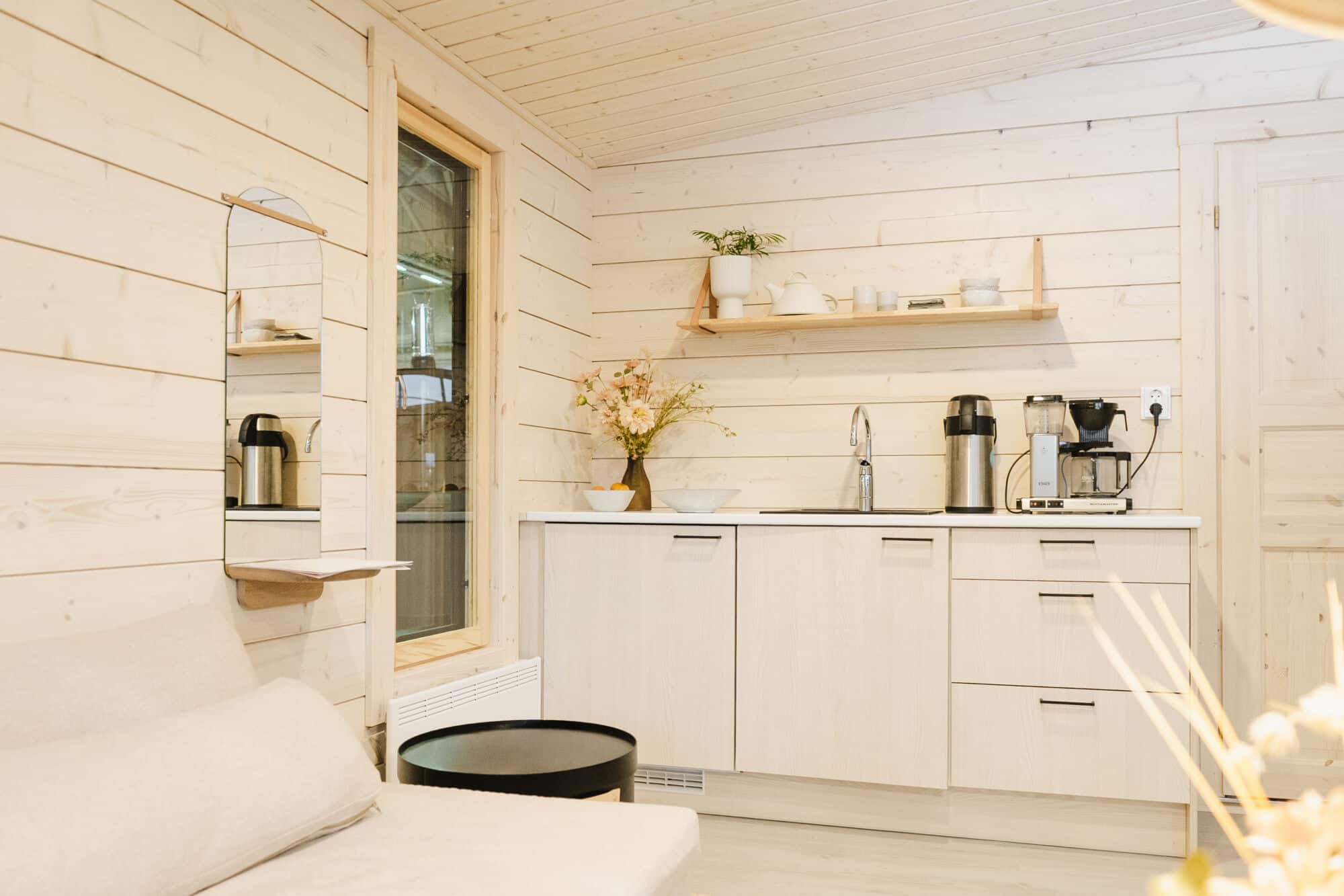 Minimalistinen keittokomero, jossa on vaaleat puukaapit, avohyllyt, työtason laitteet ja kukka-asetelma valoisassa, viihtyisässä tilassa, jossa on vaaleat puiset seinät.