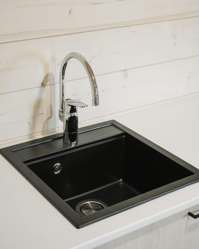 Moderni musta keittiön tiskiallas, jossa on kromihana, on asetettu valkoiseen työtasoon vaaleaa puupaneeliseinää vasten.