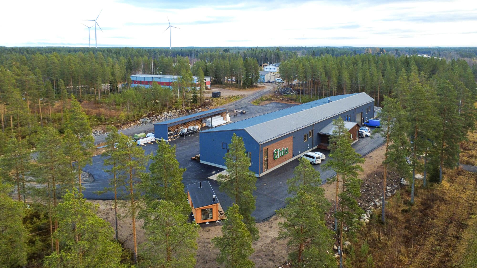 Ilmakuva puiden ympäröimästä sinisestä teollisuusrakennuksesta, jonka ulkopuolella on pysäköityjä autoja ja taustalla tuulivoimaloita - automaattinen luonnos nykyaikaisesta kestävästä suunnittelusta.