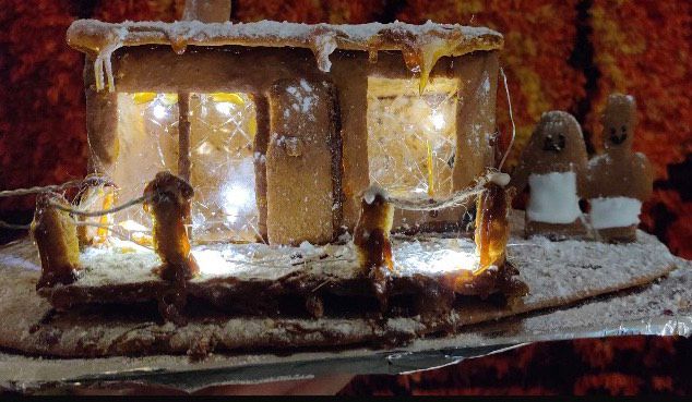 Piparkakkutalo kilpailu merkintä: piparkakkutalo koristeltu kuorrutuksella, jousivaloilla ja kaksi piparkakkuhahmoa sen vieressä, kauniisti esillä heijastavalla pinnalla.