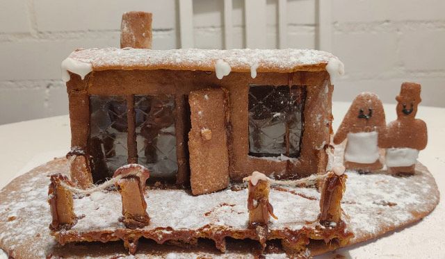 Piparkakkutalon kilpailuihin sopivassa kotitekoisessa piparkakkutalossa on kuorrutettu katto ja sen vieressä seisoo kaksi valkoisella kuorrutteella koristeltua piparkakkuhahmoa.