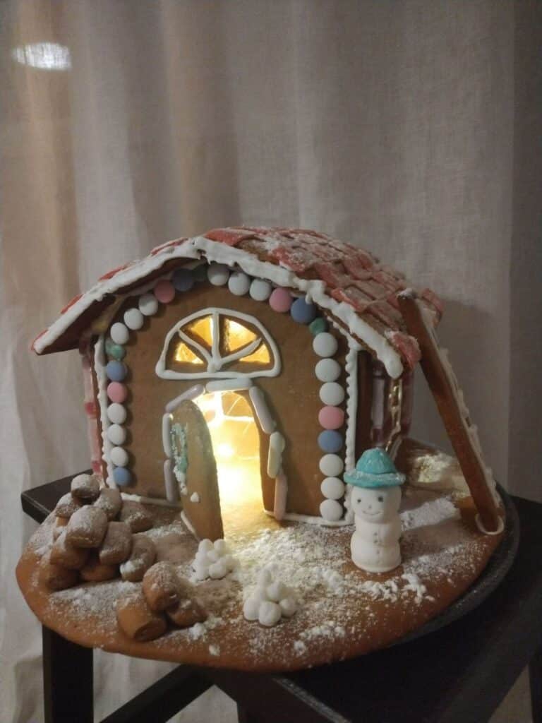 Ehtan piparkakkutalokilpailun lasten sarjan toinen voittajista. Ranskanpastilleilla koristeltu mökki, jonka edessä on halkopino ja hymyilevä lumiukko sekä lumilyhtyjä.