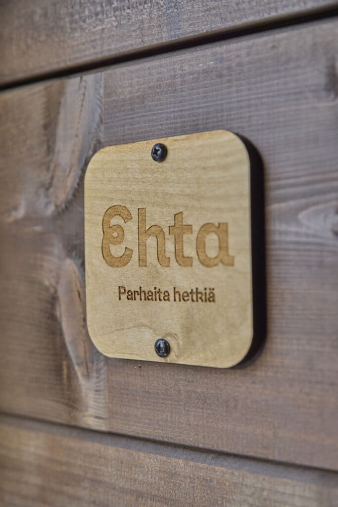 Puinen, pyöristetyillä kulmilla oleva pieni "Ehta Parhaita hetkiä" -laatta ruskeaksi maalatun hirsisen saunamökin seinässä