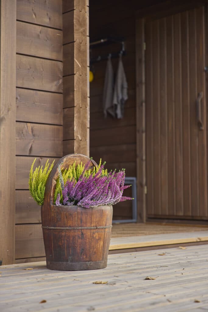 Ruskeaksi kuultomaalatun hirsirunkoisen Ehta saunamökin seinä ja terassi lähikuvassa. Terassilla kaunis vanha puuämpäri täynnä kanervia.