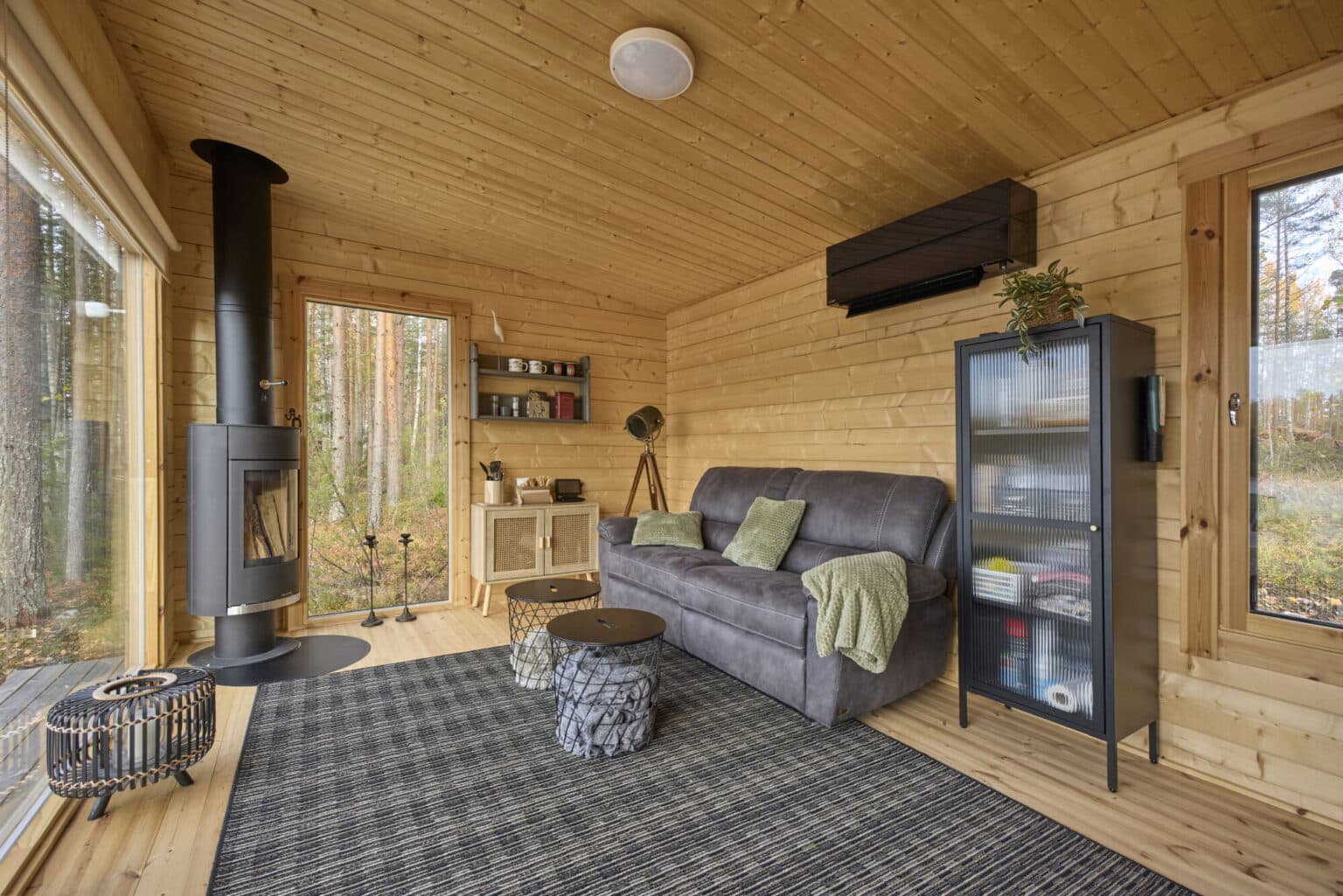 Jämsästen Ehta-saunamökin tupa on jätetty luonnollisen hirren väriseksi. Suojakäsittelyllä voi estää hirren tummumista. Sisustuksen tumman harmaat värit ja musta ilmalämpöpumppu sekä takorautainen takka luovat mukavan kontrastin modernin saunamökin tunnelmaan. Kuvassa näkyy muun muassa tumma nahkasohva, korkea lipasto lasiovilla ja mustilla kehyksillä sekä korkea maisemaikkuna, josta näkee koivikkoon.