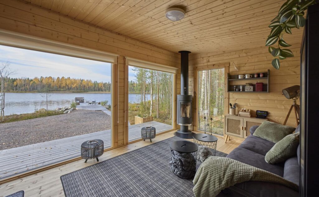 Moderni hirsipinnalla oleva Ehta saunamökin tupa. Näkymä tuvan isoista maisemaikkunoista suoraan järvelle. Nurkassa takorautainen takka ja lattialla iso, tummanharmaa kuvioitu matto.