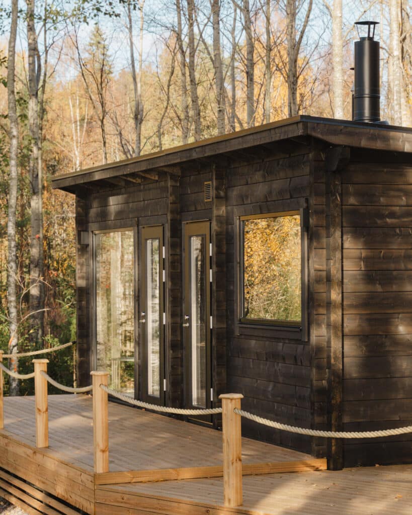 Moderni tumma puumökki, jossa on suuret ikkunat ja savupiippu ja joka sijaitsee puisella kannella metsäisellä alueella syksyllä.