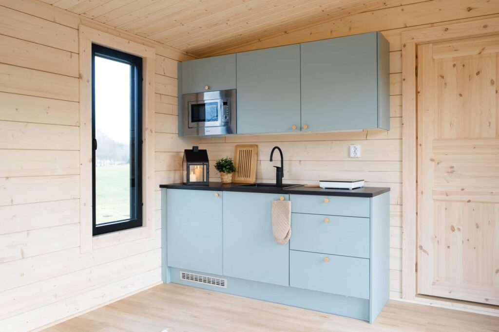 Kompakti keittiö, jossa on vaaleansiniset kaapit, musta työtaso, sisäänrakennettu mikroaaltouuni, pesuallas ja ikkuna huoneessa, jonka seinät ja lattia ovat vaaleaa puuta.