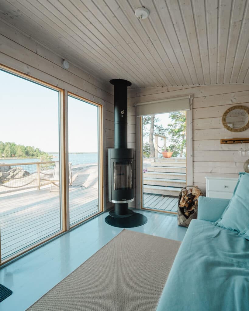 Moderni mökin sisustus, jossa on suuret ikkunat, puuhella, vaaleat puuseinät ja -katot sekä näkymä terassille, josta on näkymät veteen ja puihin.