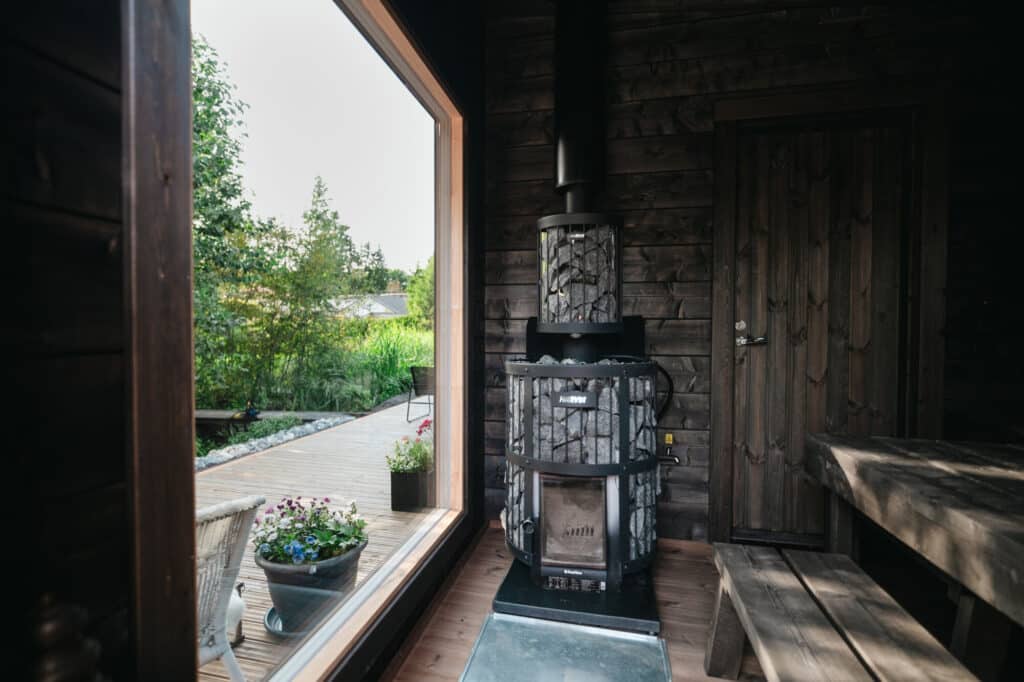 Puulämmitteinen uuni, jossa on pinottuja kiviä, sijaitsee puisessa mökkihuoneessa, jonka suuresta ikkunasta on näkymät terassille ja puutarhaan.
