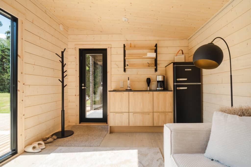 Pieni moderni mökki, jossa on vaaleat puuseinät, kompakti keittokomero, musta jääkaappi, lattiavalaisin, vaateteline ja beige sohva lasioven vieressä, josta on näkymät vehreään.