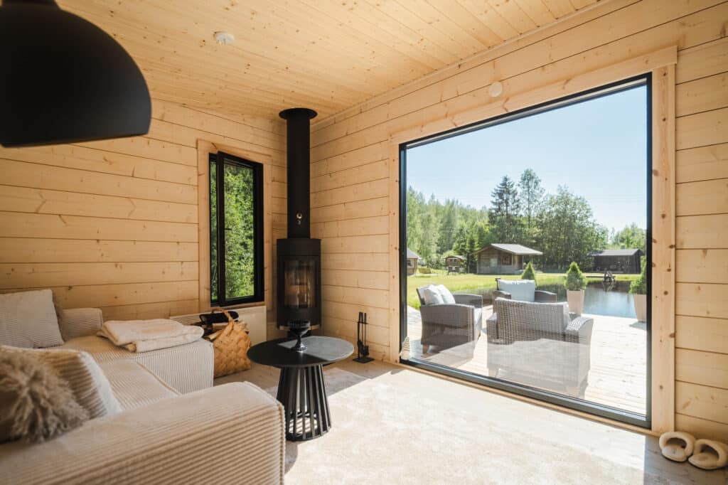 Viihtyisä, moderni huone, jossa on vaaleat puuseinät, takka ja suuri ikkuna, josta on näkymät terassille ja vehreään ulkoilmamaisemaan.