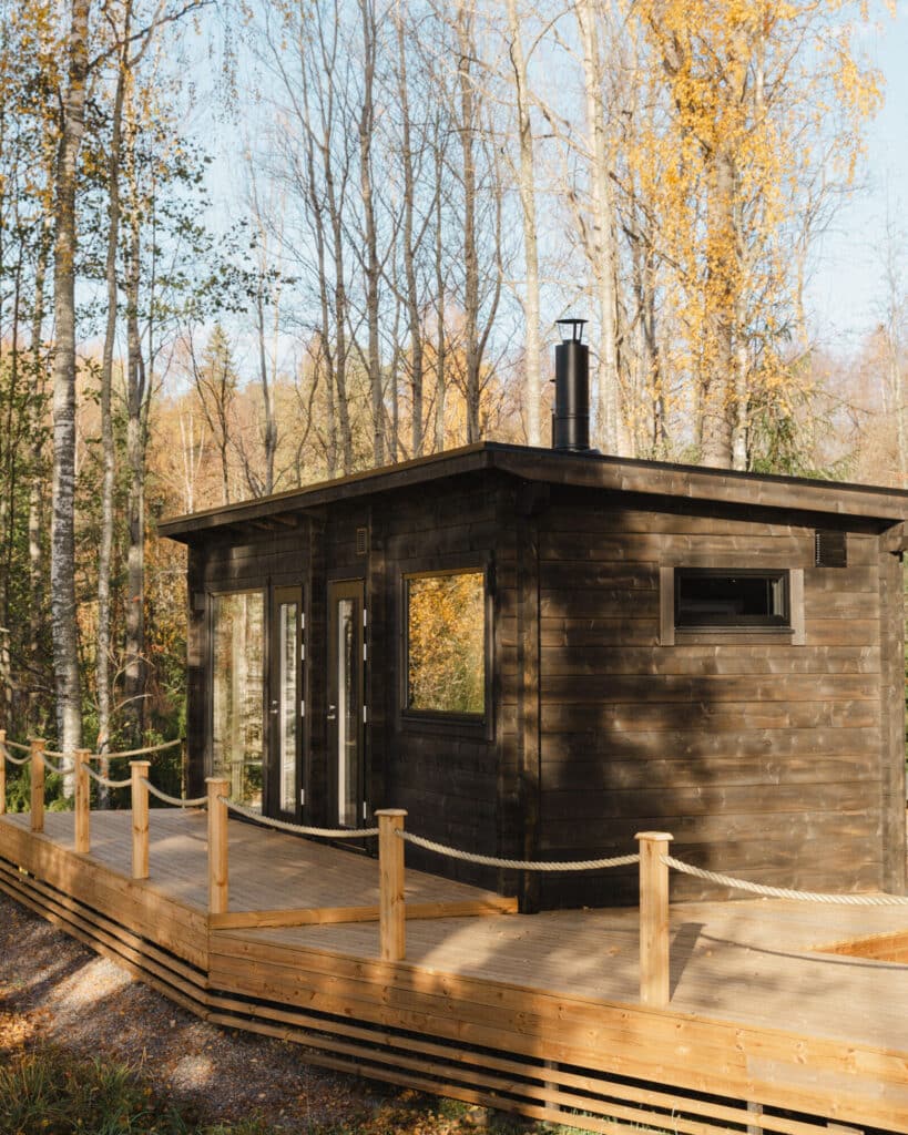 Pieni tumma puinen mökki, jossa on suuret ikkunat, sijaitsee syksyisten puiden keskellä, puisen luiskan ja köysikaiteen vieressä kirkkaan sinisen taivaan alla.