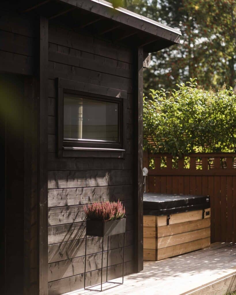 Tumma puinen vaja, jossa on pieni ikkuna, istutusastia, jossa on violetteja kukkia, ja puinen säilytyslaatikko terassilla, taustalla vehreyttä.