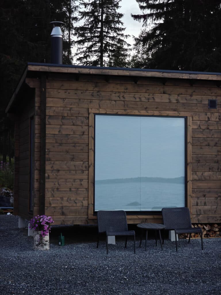 Pieni puinen mökki, jossa on suuri ikkuna, kaksi mustaa tuolia ja pieni pöytä ulkona sora-alueella, jota ympäröivät korkeat puut.