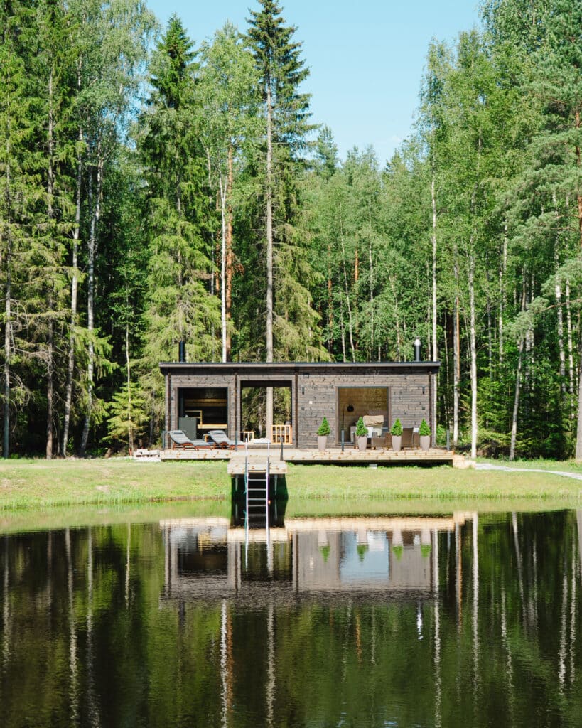 Moderni yksikerroksinen mökki, jossa on suuret ikkunat, on pienen laiturin edessä ja heijastuu rauhalliseen lampeen, jota ympäröi tiheä vihreä metsä.
