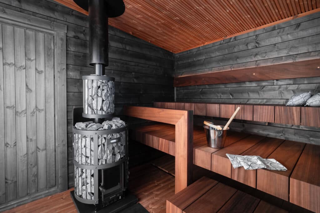 Moderni puinen saunan sisustus, jossa on tummat seinät ja penkki-istuimet, kivillä täytetty metallinen puukiuas ja penkille asetettu kauha kauhalla.
