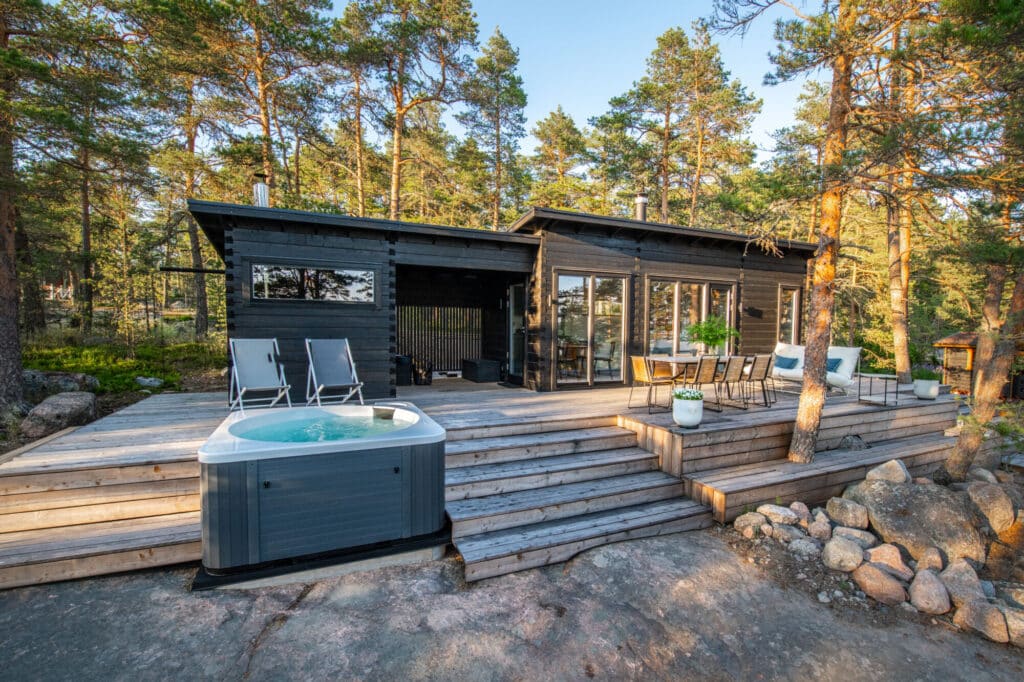 Moderni musta mökki, jossa on suuret ikkunat, tilava puinen terassi, lepotuoleja, ruukkukasveja ja poreallas, jota ympäröivät korkeat männyt ja kallioinen maasto.